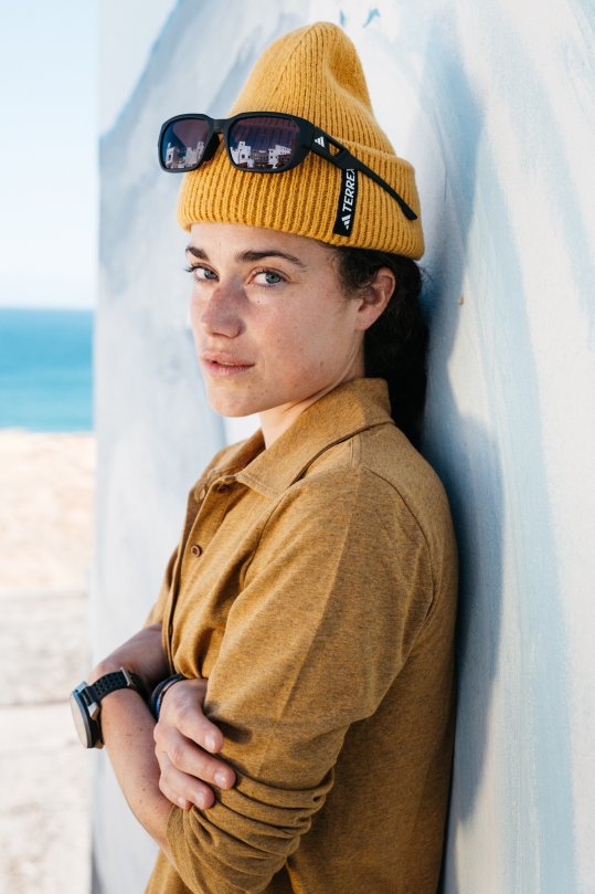 Trailsrunnerin Kimi Schreiber in gelber Kleidung an Wand gelehnt; Hintergrund: Meer und Strand