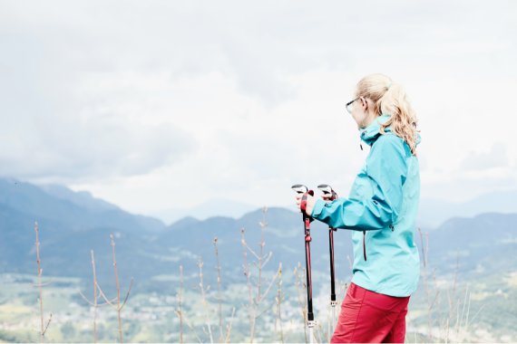 Eine Frau in Sofshelljacke macht beim Wandern eine Pause und genießt den Ausblick über die Berglandschaft