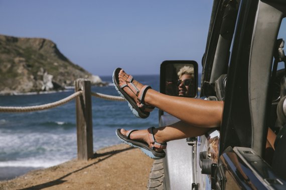 Füße mit Teva-Sandalen baumeln aus dem Autofenster am Strand