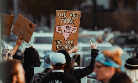 Frauen protestieren. Schild im Fokus zeigt "We are more than this - we are people"