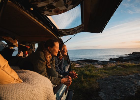 Zwei Frauen mit Kleidung von Bergans liegen in einem Camper und schauen auf das Meer bei Sonnenuntergang