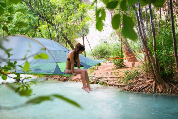 Eine junge Frau sitzt in ihrem Tentsile, das in malerischer Kulisse zwischen Bäumen über einen Bachlauf gespannt ist. 