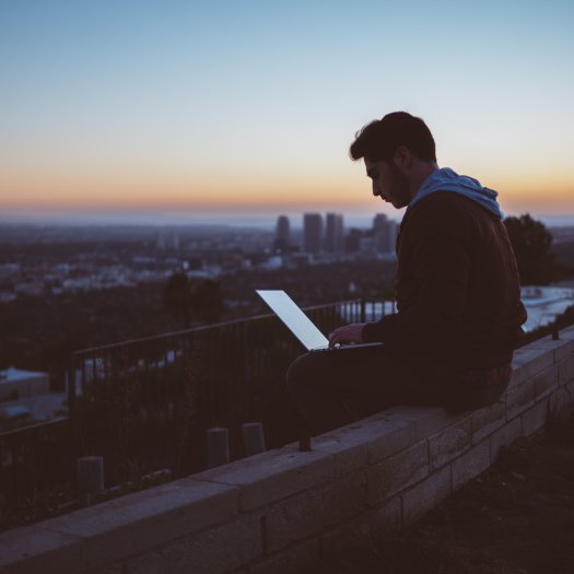 Ein Mann sitzt auf einer Mauer mit einem Laptop auf dem Schoß, im Hintergrund eine Stadt im Sonnenuntergang