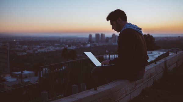Ein Mann sitzt auf einer Mauer mit einem Laptop auf dem Schoß, im Hintergrund eine Stadt im Sonnenuntergang