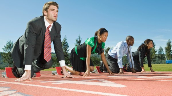 Vier Menschen stehen in Bürokleidung in Startposition auf einer Laufbahn.