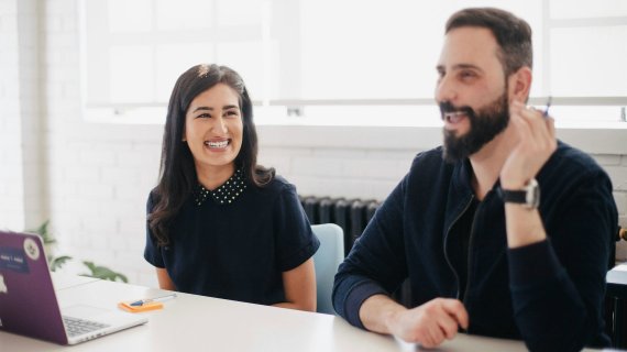 Eine Frau und ein Mann sitzen gut gelaunt an einem Bürotisch. 