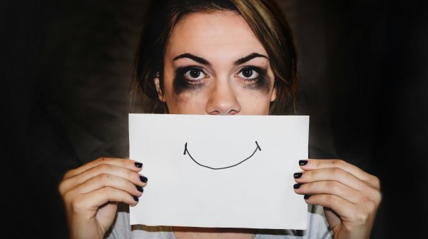 A sad woman holds a note with a happy smiley in front of her face.